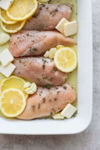 chicken breasts with lemon slices and herbs in a white baking dish.