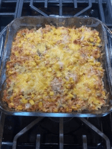 A glass baking dish filled with a chicken casserole featuring rice, corn, diced vegetables, and melted cheese on top, resting on a stovetop.