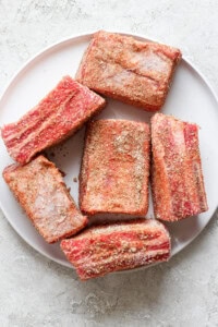 A white plate with six seasoned raw beef short ribs on a light gray surface.