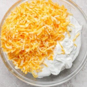 A clear glass bowl filled with shredded yellow and white cheese on top of a mound of white, creamy substance on a light-colored surface.