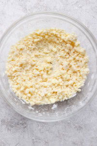 A clear glass bowl containing a crumbly mixture of flour and butter, sitting on a textured gray surface.