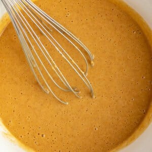 A metal whisk in a white bowl containing smooth, light brown cake batter.