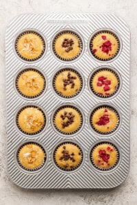 A muffin tray with 12 muffins, topped with almonds, chocolate chips, and raspberries.