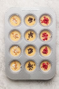 Muffin batter in a tray with toppings including coconut, chocolate chips, and raspberries.