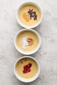 Three bowls of batter are topped with different ingredients: chocolate chips and peanut butter, shredded coconut and nuts, and red berries, arranged vertically on a textured surface.