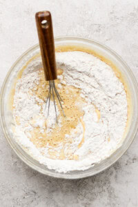 A glass bowl with a wooden-handled whisk mixing flour into a batter on a textured surface.