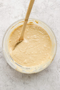 A glass bowl filled with creamy batter and a wooden spoon rests in it on a textured white surface.