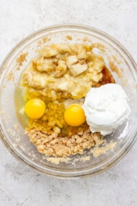 A glass bowl containing mashed bananas, two eggs, yogurt, brown sugar, and a liquid ingredient.