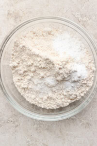 A clear glass bowl containing a mixture of flour and baking powder on a textured beige surface.
