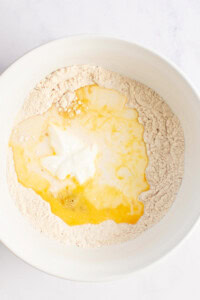 A white bowl containing flour with a well in the center holding cracked eggs and yogurt.