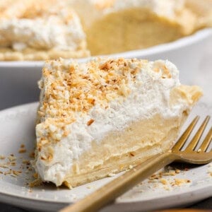 A slice of coconut cream pie on a white plate with a gold fork, topped with whipped cream and toasted coconut flakes.