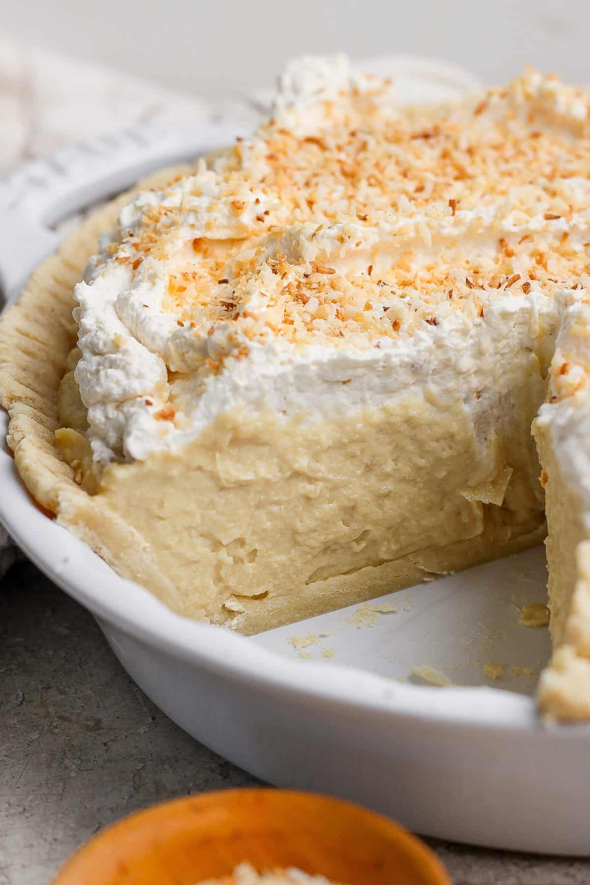 A creamy pie with a slice removed, topped with whipped cream and toasted coconut flakes, in a white pie dish.