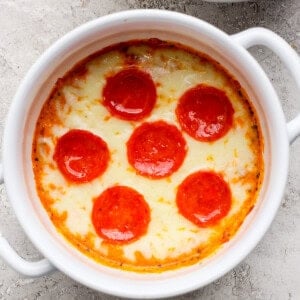 Baked dish topped with melted cheese and six evenly spaced pepperoni slices in a white ramekin on a textured surface.