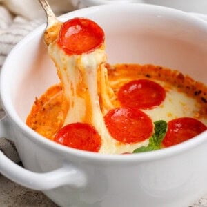 A spoon lifts cheesy pepperoni pizza dip from a white bowl. Fresh basil leaves are visible nearby.