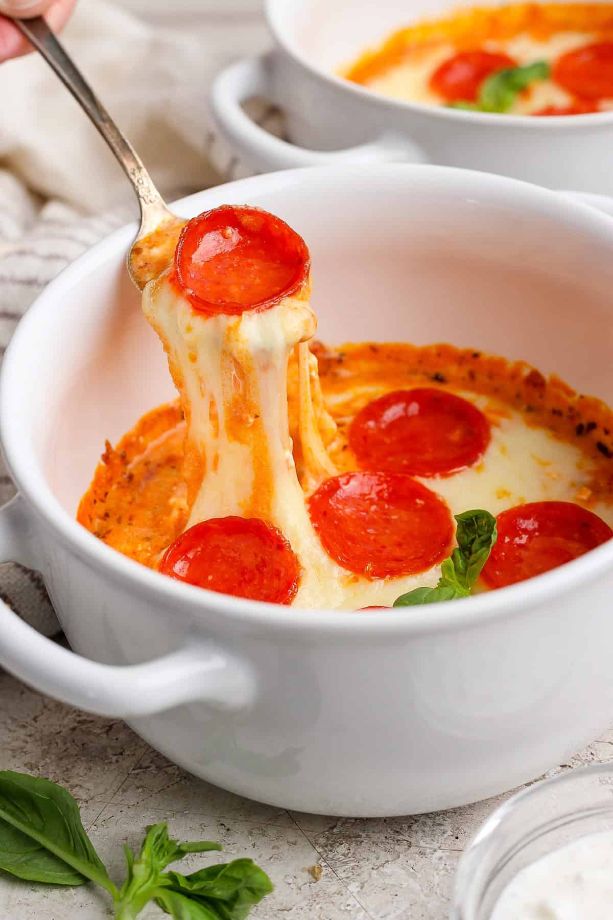 A spoon lifts cheesy pepperoni pizza dip from a white bowl. Fresh basil leaves are visible nearby.