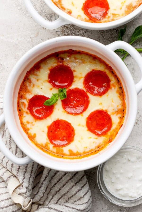 A white bowl filled with baked pepperoni and cheese dip, garnished with a basil leaf, placed on a cloth next to a small bowl of dipping sauce.