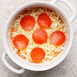 A round white dish containing shredded cheese and six slices of pepperoni on a textured surface.