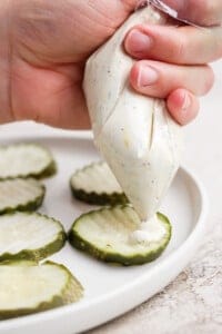 A hand squeezes creamy sauce from a piping bag onto crinkle-cut pickle slices arranged on a white plate.