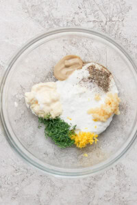 Glass bowl with Greek yogurt, mayo, Dijon mustard, minced garlic, chopped dill, lemon zest, black pepper, and salt on a textured surface.