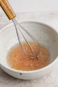 A metal whisk rests in a white bowl filled with a light brown liquid, possibly a vinaigrette or sauce, set on a textured surface.