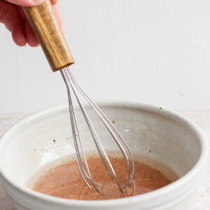 A hand using a wooden-handled whisk to stir liquid in a ceramic bowl.