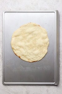 A round, uncooked pizza dough is placed on a metal baking sheet with a textured surface.