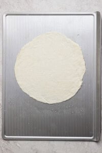 A round, uncooked pizza dough is placed on a textured metal baking sheet.