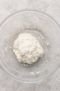 A ball of dough in a clear glass bowl on a light textured surface.