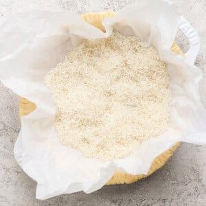 A bowl lined with parchment paper filled with uncooked white rice, placed on a textured surface.