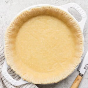 Unbaked pie crust in a white ceramic pie dish with a striped cloth and a knife beside it on a textured surface.