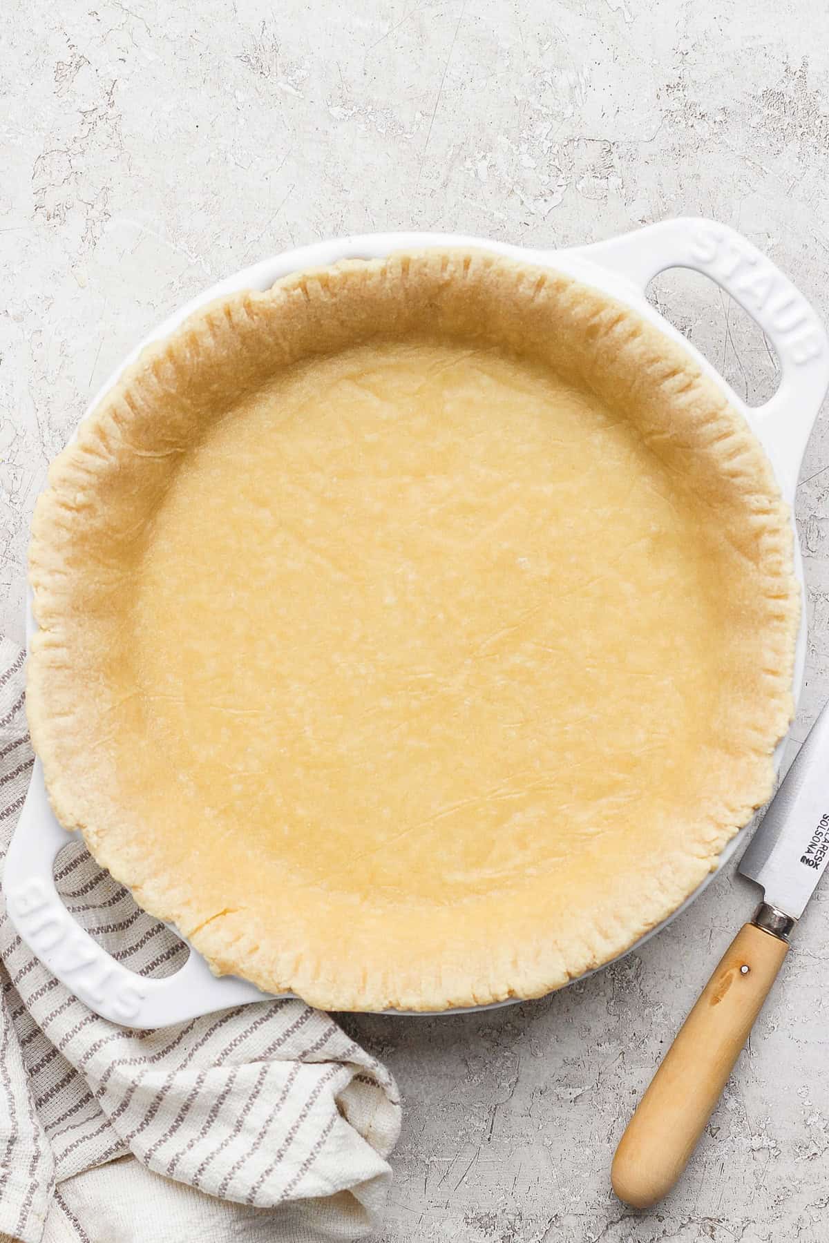 Unbaked pie crust in a white pie dish on a textured surface with a knife and striped cloth nearby.