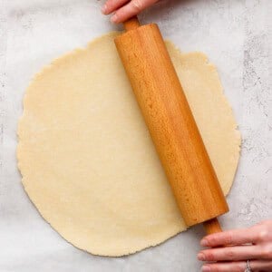 Hands rolling out dough on parchment paper with a wooden rolling pin.