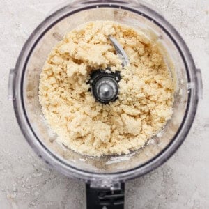 A food processor containing crumbly dough mixture, placed on a textured gray surface.