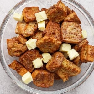 A glass bowl filled with croutons and small cubes of butter on a light gray textured background.