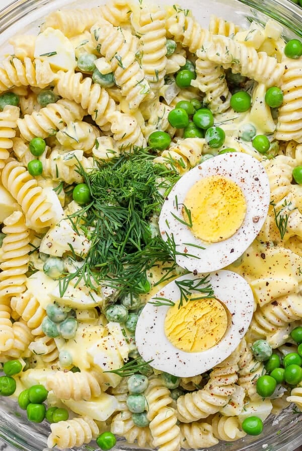 A bowl of fusilli pasta salad with green peas, chopped dill, and sliced hard-boiled egg halves on top.