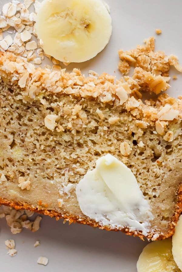 Slice of banana bread with butter and oats, surrounded by banana slices, on a white plate.