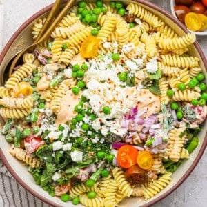 Bowl of pasta salad with rotini, peas, cherry tomatoes, crumbled cheese, greens, and a creamy dressing, garnished with chopped onions and red pepper flakes.