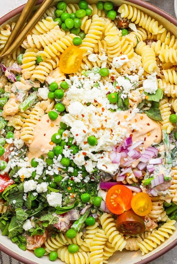 Bowl of pasta salad with rotini, peas, cherry tomatoes, crumbled cheese, greens, and a creamy dressing, garnished with chopped onions and red pepper flakes.