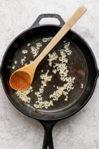 A cast iron skillet with sautéing minced garlic in oil, accompanied by a wooden spoon.
