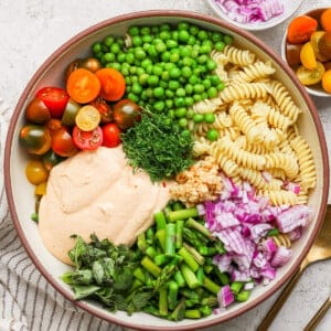 A bowl with rotini pasta, cherry tomatoes, peas, chopped red onion, green beans, fresh herbs, and a creamy sauce. A small bowl of onions and cutlery are beside it.