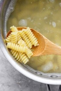A wooden spoon holding cooked rotini pasta above a pot of boiling water.