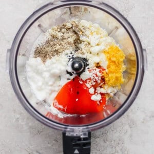 Ingredients including pepper, feta cheese, cottage cheese, lemon zest, and red peppers in a food processor, ready to be blended.
