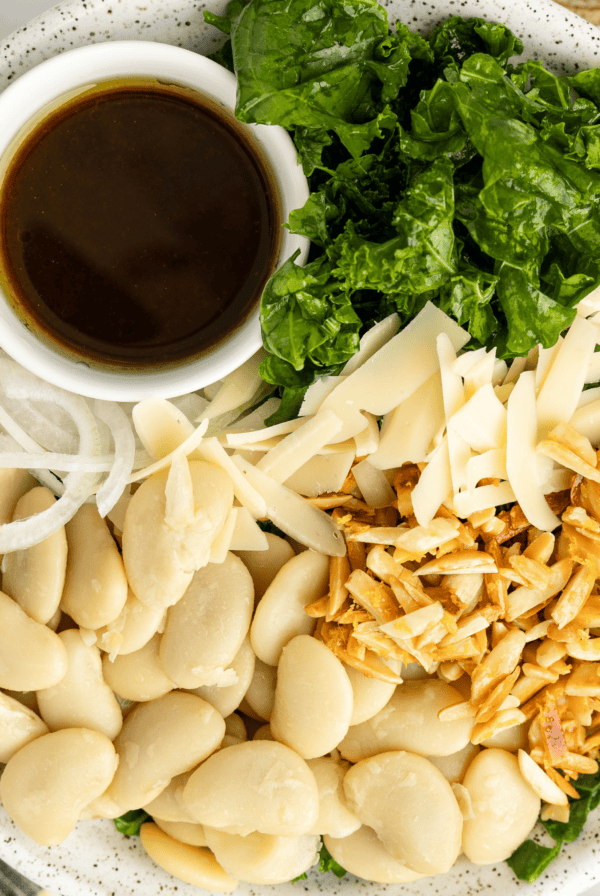 A plate with butter beans, sliced onions, kale, shredded cheese, toasted slivered almonds, and a small cup of dark dressing.