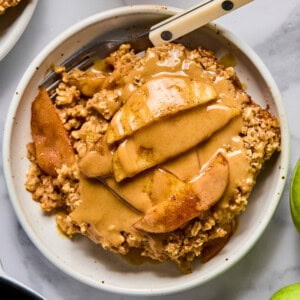A bowl of apple crisp topped with sliced apples and a generous drizzle of nut butter, with a fork resting in the bowl.