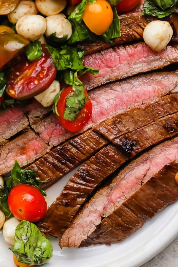 Sliced medium-rare steak served with a salad of cherry tomatoes, mozzarella balls, and fresh basil on a white plate.