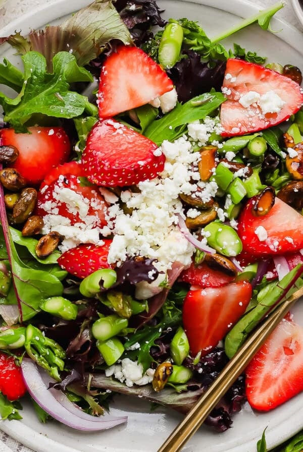 A plate of mixed green salad topped with sliced strawberries, feta cheese, red onion, pistachios, asparagus, and a gold fork on the side.