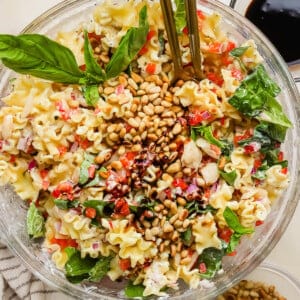A glass bowl of pasta salad with spinach, pine nuts, red onion, and chopped vegetables, garnished with basil leaves and served with dressing on the side.