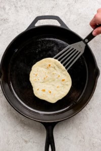 A hand uses a spatula to lift a tortilla cooking in a black cast iron skillet on a light-colored surface.