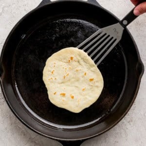 A hand uses a spatula to lift a tortilla cooking in a black cast iron skillet on a light-colored surface.