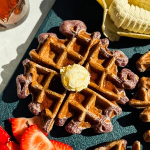 A waffle topped with a dollop of butter is surrounded by sliced bananas and strawberries on a dark surface.